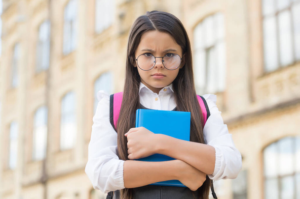 Unhappy little kid in eyeglasses hold study book in schoolyard outdoors, back to school - Foto, Imagem