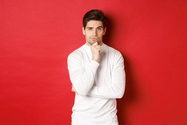 Kép a figyelmes jóképű férfi, hogy feltevés, gondolkodás és a kamera, álló fehér pulóver vörös háttér - Fotó, kép