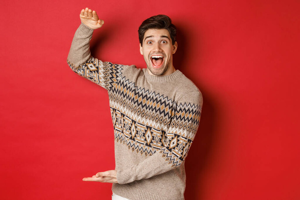 Portrét vzrušeného a šťastného pohledného muže ve vánočním svetru, ukazuje něco velkého, drží velký dárek na dovolenou, stojí nad červeným pozadím - Fotografie, Obrázek