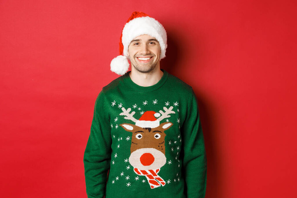 Pojęcie ferii zimowych, Bożego Narodzenia i stylu życia. Przystojny facet z włosia, w kapeluszu Mikołaja i zielonym swetrze, uśmiechnięty radosny, świętujący Nowy Rok, stojący na czerwonym tle - Zdjęcie, obraz