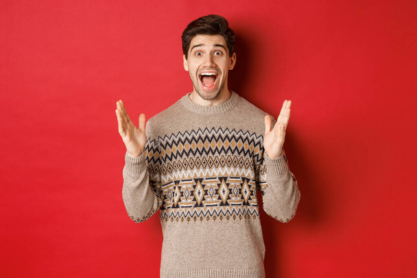 Portrait of surprised and happy handsome man, wearing christmas sweater, looking amazed, celebrating new year, standing over red background - Photo, Image