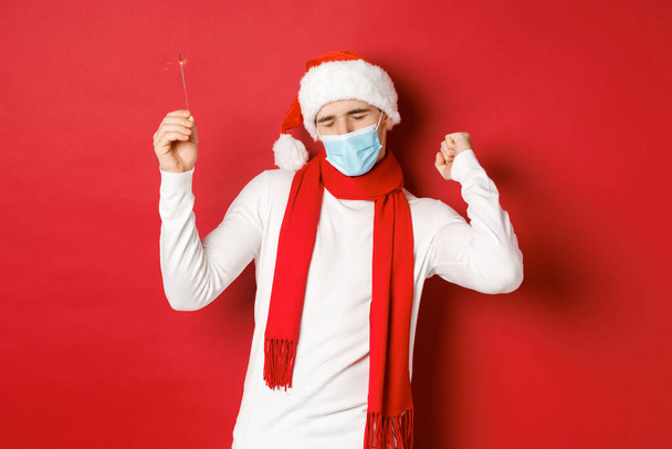 Концепция ковида-19, Рождества и праздников во время пандемии. Счастливчик празднует Новый год на вечеринке, носит медицинскую маску и шляпу Санты, танцует с блеском на красном фоне - Фото, изображение