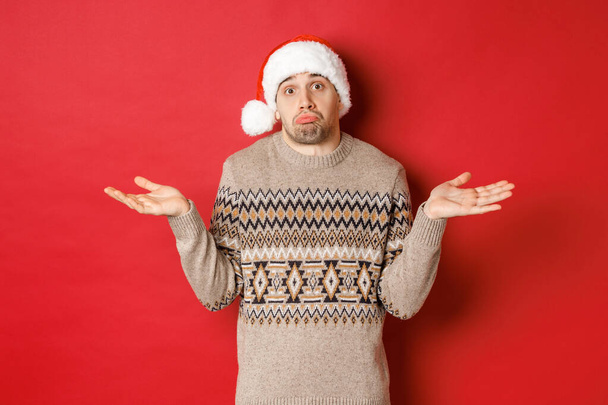 Εικόνα του ανίδεου νέου άνδρα με πουλόβερ και καπέλο santa, shrugging και αναζητούν άγνοια, dont ξέρει τι να αγοράσει για τις διακοπές του νέου έτους, στέκεται πάνω από το κόκκινο φόντο - Φωτογραφία, εικόνα