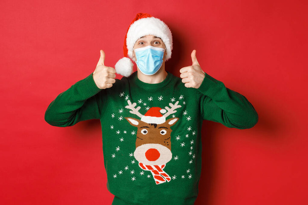 Concept of new year, covid-19 and social distancing. Cheerful handsome man in santa hat and medical mask, showing thumbs-up, enjoying christmas party, standing over red background - Zdjęcie, obraz