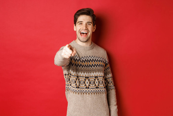 Концепція святкування Різдва, зима і спосіб життя. Щасливий привабливий чоловік в різдвяному светрі, бажаючи щасливих свят і вказуючи на камеру, стоїть на червоному тлі
 - Фото, зображення
