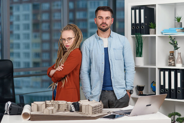 Handsome professional young engineer stands back to back with pretty female colleague with dreadlocks at design bureau. - 写真・画像