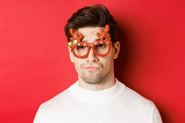 Kış tatili, Noel ve kutlama kavramı. Parti gözlüğü takan, sırıtan, eğlenmemiş, kırmızı arka planda duran, şüpheli yakışıklı adamın yakın çekimi. - Fotoğraf, Görsel