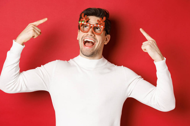 Handsome happy guy in white sweater, pointing at his christmas party glasses, celebrating new year and having fun, standing over red background - Photo, Image
