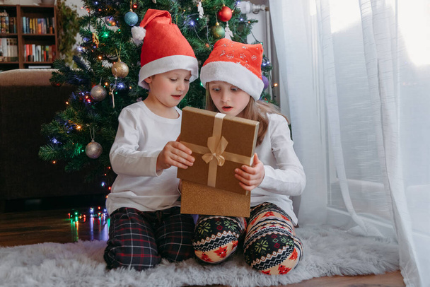 Um menino e uma menina de chapéus de Natal abrem uma caixa de presente debaixo de uma árvore de Natal. Celebração, receber presentes, alegria das crianças. Milagre, presente - Foto, Imagem