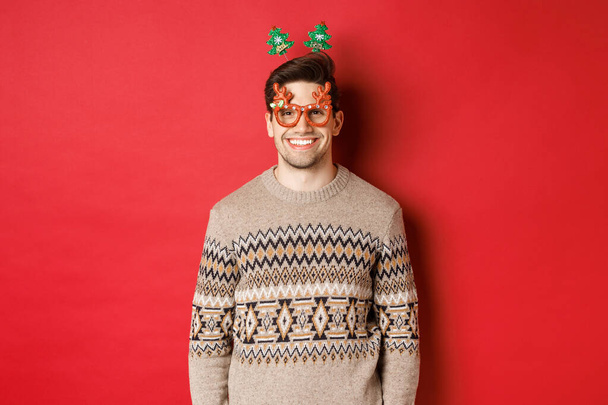 Retrato de cara barbudo bonito em óculos de festa e camisola de inverno, sorrindo satisfeito, celebrando o ano novo, desejando Feliz Natal, de pé sobre fundo vermelho - Foto, Imagem