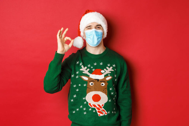 Concept of new year, covid-19 and social distancing. Cheerful man in medical mask and santa hat, showing okay sign, recommending something good, standing over red background - 写真・画像