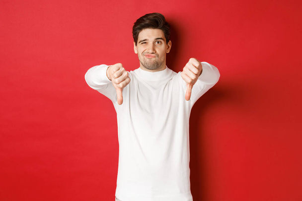 Portrét zklamaného skeptika, v bílém svetru, ukazující palce dolů, aby nesouhlasil, vyjadřující odpor, stojící nad červeným pozadím - Fotografie, Obrázek