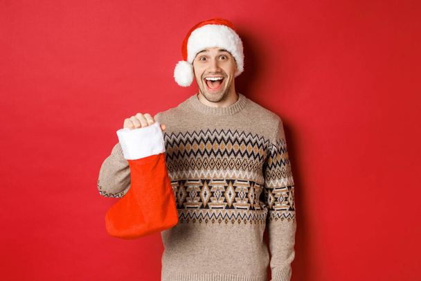 Begrip wintervakantie, nieuwjaar en viering. Vrolijke en verraste volwassen man ontvangt snoepjes op Sint Nicolaas dag in rode kous, staande verbaasd over rode achtergrond - Foto, afbeelding