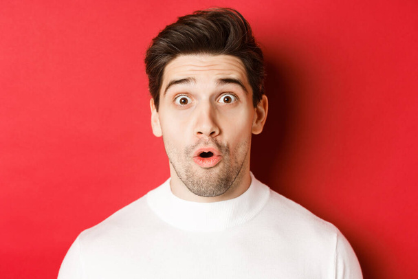 Close-up of surprised handsome man in white sweater, open mouth and looking wondered, hear amazing news, standing over red background - Photo, Image