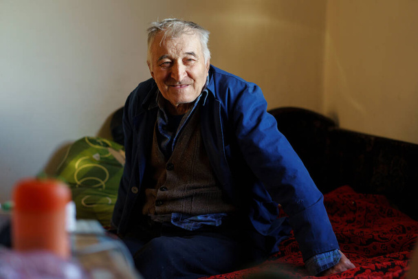 Starší běloch jeden starý důchodce sedí na posteli v pokoji v pečovatelském domě sám ve dne šedé vlasy muž dívá na stranu usmívá - Fotografie, Obrázek