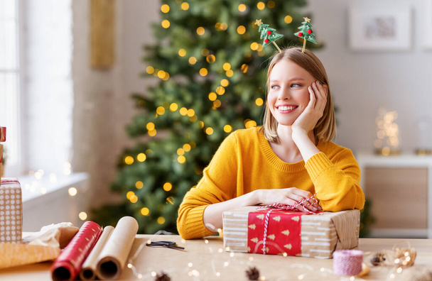 Glückliche junge Frau mit Geschenkbox schaut weg und träumt, während sie am Weihnachtstag am Tisch im gemütlichen Zimmer hom sitzt - Foto, Bild