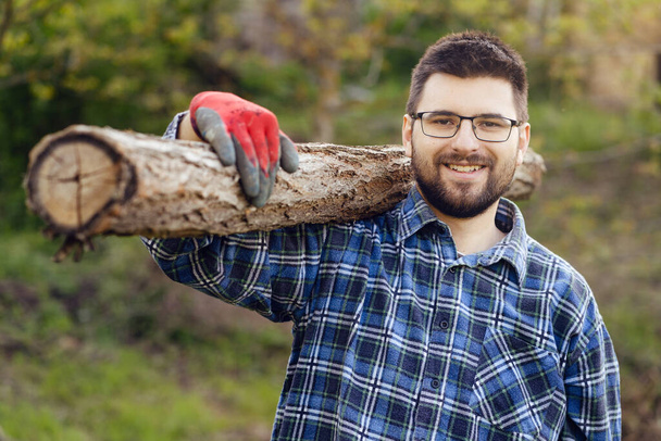 Ritratto di giovane uomo caucasico adulto taglialegna operaio che indossa camicia barba e guanti da lavoro protettivi che tiene un tronco di tronco di tronco sulla spalla in cortile all'aperto di giorno sorridente vista frontale - Foto, immagini
