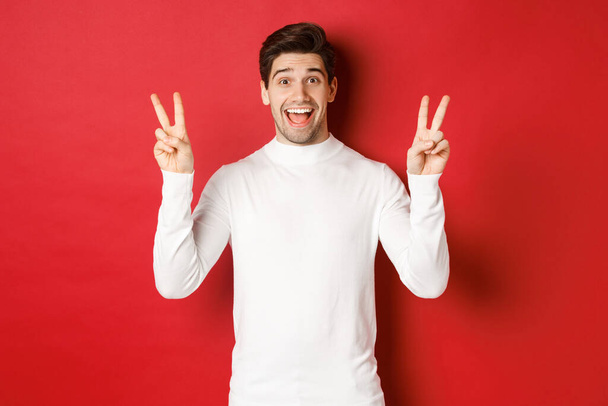 Concept of winter holidays, christmas and lifestyle. Handsome funny guy in white sweater, showing peace signs and smiling happy, standing against red background - Photo, Image