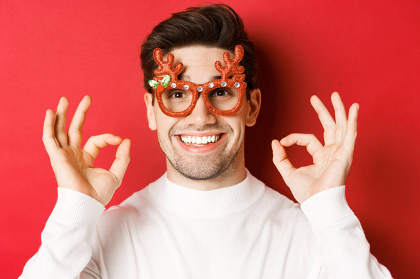 Conceito de férias de inverno, Natal e celebração. Close-up de homem atraente em óculos de festa, mostrando sinais ok e sorrindo, louvar algo bom, fundo vermelho - Foto, Imagem