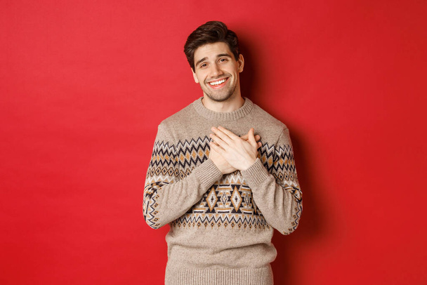 Porträt eines gerührten und glücklichen gutaussehenden Kerls, der Neujahrsgeschenk erhält, die Hände auf dem Herzen hält und lächelt, Danke sagt, im Weihnachtspullover vor rotem Hintergrund steht - Foto, Bild