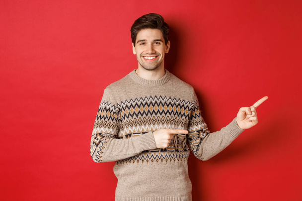 Wizerunek atrakcyjnego uśmiechniętego mężczyzny, noszącego świąteczny sweter, wytykającego palcami w prawo i pokazującego noworoczną reklamę, stojącego na czerwonym tle - Zdjęcie, obraz
