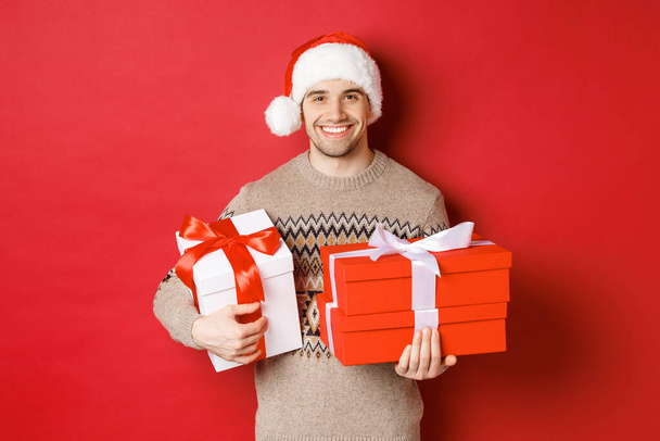 Concept of winter holidays, new year and celebration. Portrait of handsome man in santa hat and sweater, holding boxes with christmas presents and smiling, prepared gifts, red background - Fotó, kép