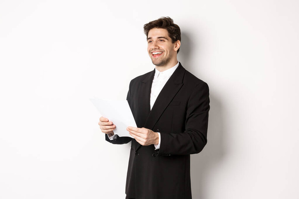 Image of handsome businessman in suit, holding documents and smiling, standing against white background - Фото, изображение