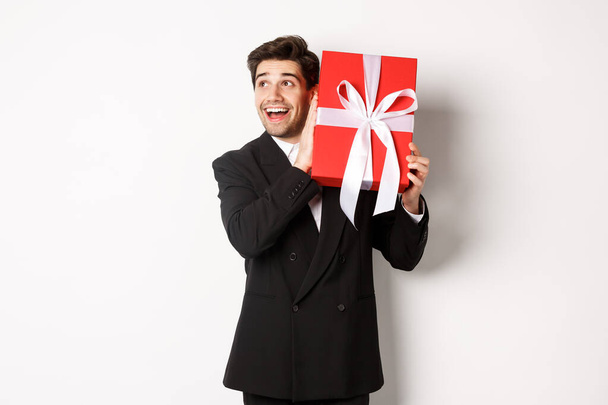 Obrázek pohledný zasněný chlap v černém obleku, třesoucí se box s dárkem divit, co je uvnitř, stojí na bílém pozadí šťastný - Fotografie, Obrázek