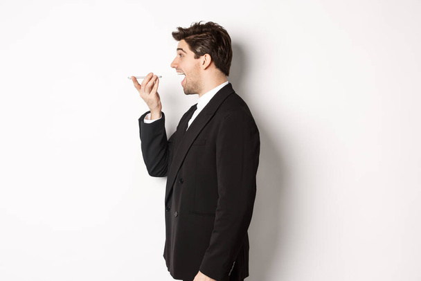 Profile shot of handsome businessman in black suit talking on speakerphone, smiling and looking happy, recording voice message, standing over white background - Photo, Image