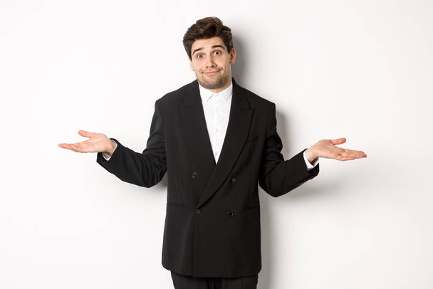 Image of handsome confused man in black suit, shrugging and looking clueless, dont know anything, standing over white background - Photo, Image
