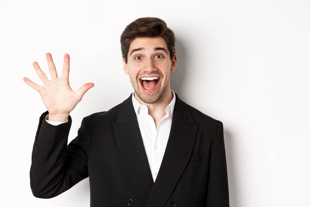 Close-up of handsome businessman in black suit, smiling amazed, showing number five, standing over white background - Foto, Imagen
