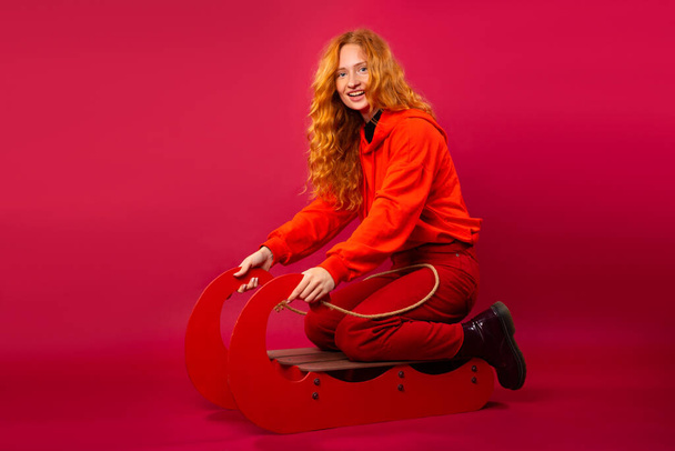 Satisfied young red-haired girl on a sled. Funny Face. Winter fun concept. Photo on a red background. High quality photo - Photo, image
