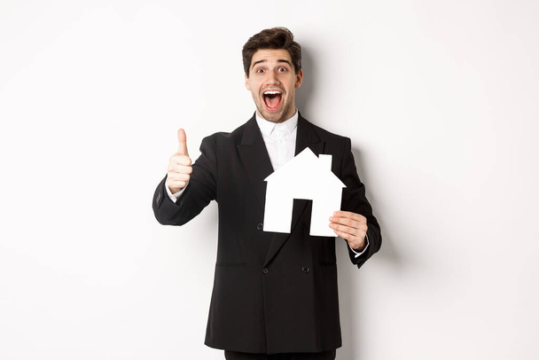 Portrait of excited handsome man looking for a home, showing house and thumb-up, recommending real estate agency, standing against white background - Photo, Image
