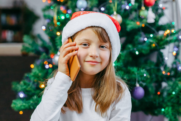 Online Christmas greetings. A close-up portrait of a cute girl in a New Years hat with a mobile phone. The child uses gadgets to congratulate family and friends - Foto, Imagen