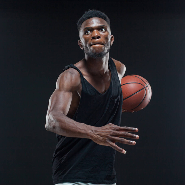 Portrait of afro american male basketball player playing with a ball over black background. Fit young man in sportswear holding basketball - Foto, Imagen