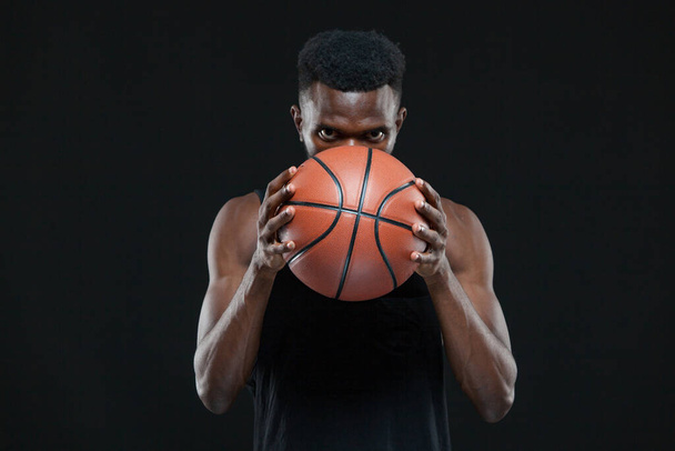 Close up front view shot of afro american male basketball player holding a ball in front of him over black background - Photo, image