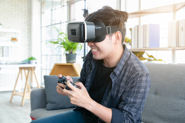 Attractive young man wearing virtual reality headset or 3d glasses sitting on sofa at home. - 写真・画像