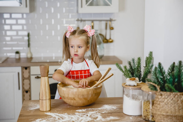 Four-year-old blonde girl in a white t-shirt and red apron kneads dough at home in the kitchen at a wooden table - Foto, Imagem