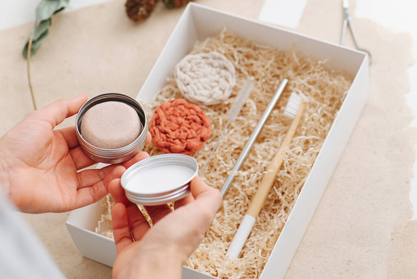 Female hand picking setting powder form box with multi-use eco-friendly items. It also contains wooden brush, steel straw and woven makeup remover discs on a filling paper. - Foto, imagen