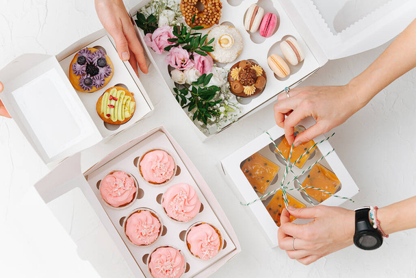 Female hands beautifully and carefully arranging colorful sweets in white windowed boxes. Zefir, cookies, macarons and cakes decorated with branches and flowers. Top view. - Foto, Bild