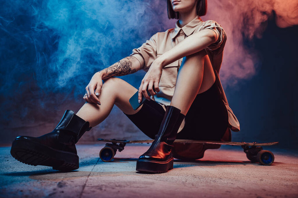 Öltözött trendi és egyszerű ruházat lány rövid frizura és tetoválás pózol ül a korcsolya és gazdaság okostelefon füstös háttér. - Fotó, kép