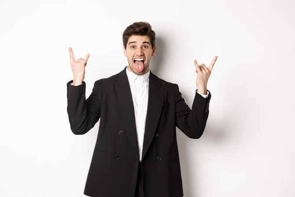 Portrait of happy attractive guy having fun at party, wearing black suit, showing rock-n-roll sign and tongue, standing excited against white background - Photo, image