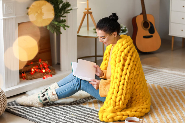 Beautiful young woman with warm plaid reading book at home - Foto, Bild