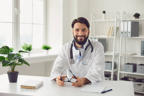 Happy bearded doctor sitting at table in medical office. - Foto, Bild