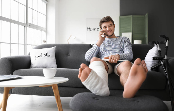 Young man with broken leg talking by phone at home - Foto, Bild