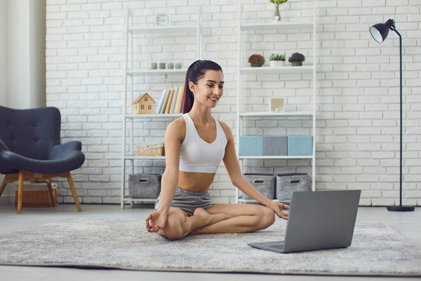 Girl sitting on carpet in lotus position and practicing yoga during online lesson on laptop at home - Φωτογραφία, εικόνα