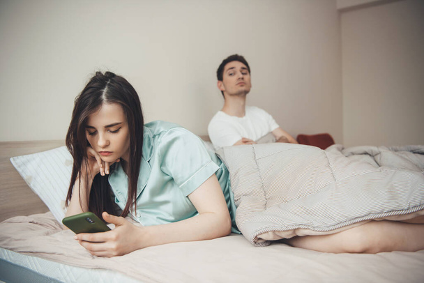 Ο ζηλιάρης καυκάσιος προσπαθεί να δει σε ποιον πληκτρολογεί η κοπέλα του, χρησιμοποιώντας ένα τηλέφωνο στο κρεβάτι. - Φωτογραφία, εικόνα