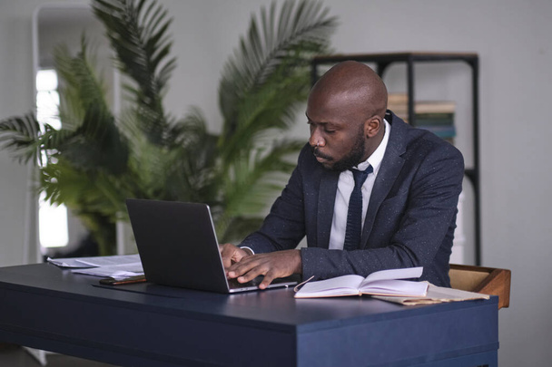 Młody afrykański kierownik czyta dokumenty i pracuje nad laptopem siedząc przy biurku w biurze. miękkie skupienie - Zdjęcie, obraz