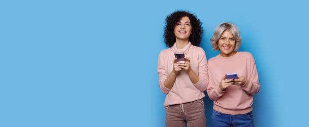 Charming blonde lady with her curly haired friend are chatting on a blue studio wall near free space smiling at camera - Photo, Image