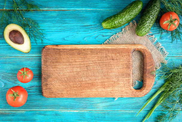 Fresh vegetables, wooden board and knife on blue background. Tomatoes, cucumbers, avocado, dill and green onion.  - Photo, Image